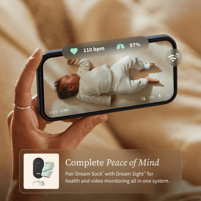 Hand holding a smartphone displaying a baby monitor app with a baby lying on a bed.