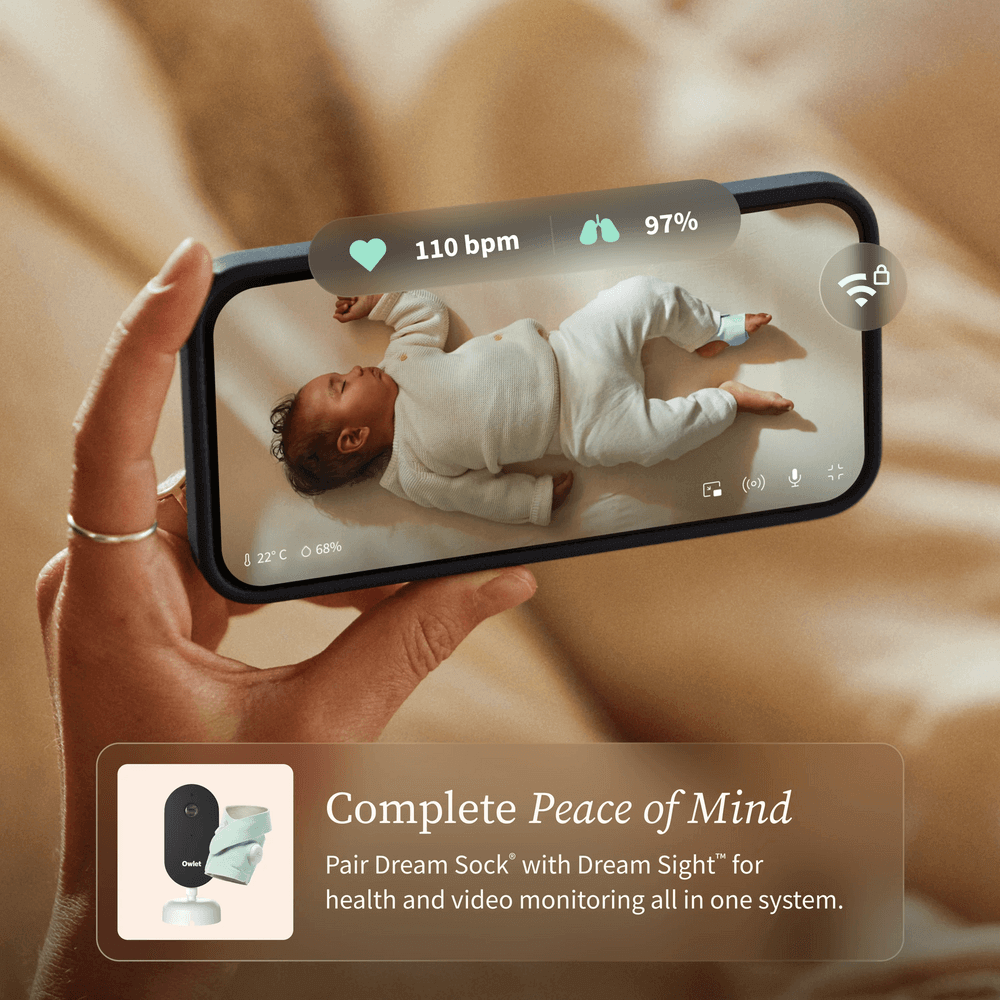 Hand holding a smartphone displaying a baby monitor app with a baby lying on a bed.
