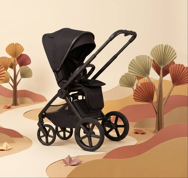 A black stroller with a canopy and push handle is shown in a stylized, minimalist paper-cut landscape scene with beige hills, winding paths, and decorative trees in warm tan, brown, and muted orange tones.