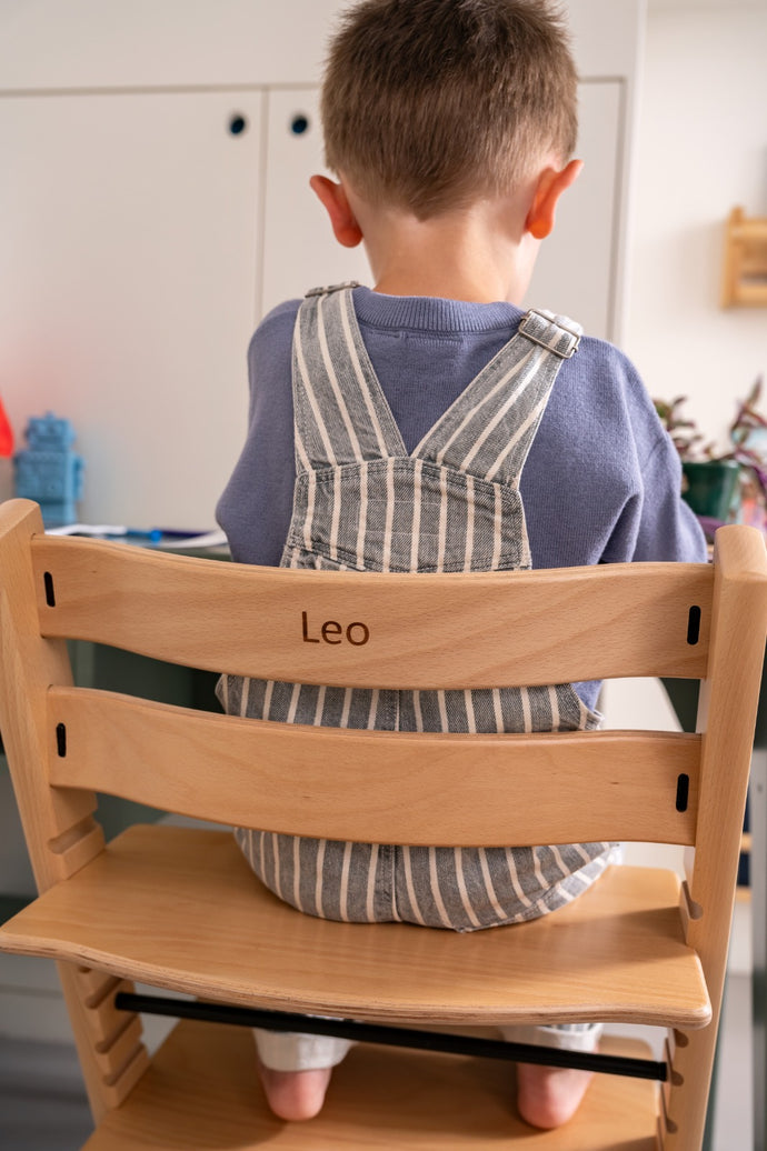 Child sitting on a wooden chair with 'Leo' on the backrest in a kitchen setting.
