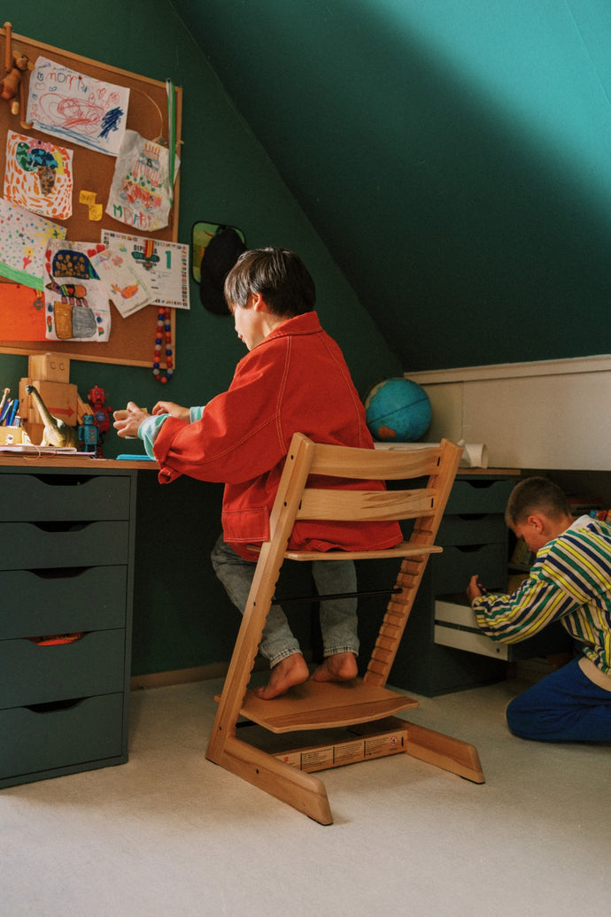 Two children in a room with a desk and high chair, one child is sitting on the high chair and the other is on the floor.
